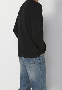 Homme portant un pull noir à manches longues côtelé et un jean bleu délavé, debout les mains dans les poches, dos à l'appareil photo.