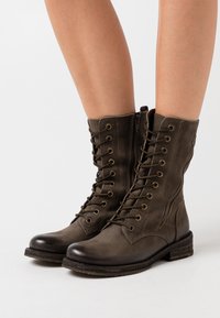 Botas de estilo militar con cordones en cuero oliva oscuro. Presentan una punta redondeada, ojetes de metal, detalles de costura y una suela texturizada.