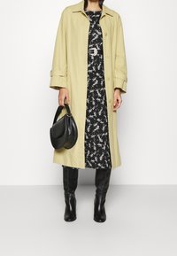 Manteau trench beige clair avec un large col et des boutons. Robe noire à fleurs en dessous, associée à des bottes noires jusqu'aux genoux et un sac à main noir texturé.