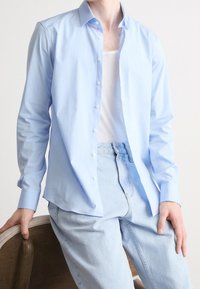 Camisa de botones azul claro con mangas largas, hecha de tela lisa. Se lleva sobre una camiseta blanca sin mangas y se combina con jeans de mezclilla clara.