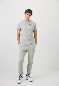 Muškarac stoji u svijetlosivoj majici i odgovarajućim trenerkama, s crno-bijelim tenisicama, na blijedoj bijeloj pozadini.