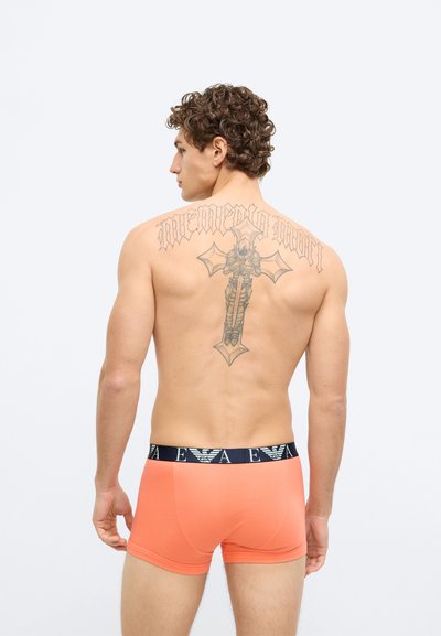 Cuecas boxer laranja com cintura azul marinho com logótipo. O modelo mostra uma tatuagem nas costas e tem cabelo encaracolado.