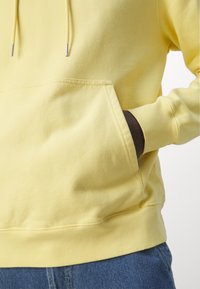 Felpa gialla in cotone con tasca frontale e cordini. Presenta polsini e orlo a coste, indossata infilata in un jeans blu.
