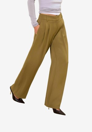 Personne portant un pantalon large olive, des escarpins noirs à bout pointu et une chemise à manches longues gris clair, une main dans la poche.