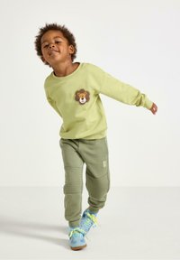 Grøn sweatshirt med et løveprint og matchende grøn sweatpants med teksturerede detaljer. Blå sko med gule snørebånd.