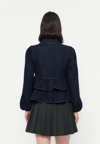 Denim peplumjacket in donkerblauw met opgezette mouwen, voorzien van een gelaagd zoom en zichtbare stiksels. Gecombineerd met een geplooide rok.