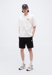 Νέος άνδρας που στέκεται με τα χέρια στις τσέπες, φορώντας λευκό μπλουζάκι Calvin Klein, μαύρο σορτς, μαύρο καπέλο, λευκές κάλτσες και λευκά αθλητικά παπούτσια.