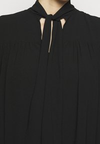 Blouse noire avec un col à nouer, présentant des plis autour de l'encolure et des découpes en forme de trou de clé. Tissu lisse avec une texture légère.