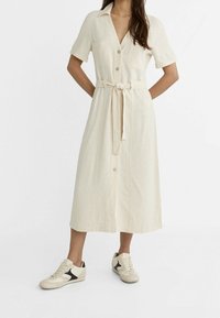 Robe chemise en lin beige avec des manches courtes, fermée par des boutons sur le devant, longueur genou, et une ceinture à nouer. Associée à des baskets crème avec des accents noirs.