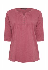 Vaaleanpunainen lyhythihainen paita, jossa on pyöreä pääntie, napillinen etulerssi ja pystyröyhelöitä lisätekstuurin tuomiseksi.