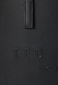 Sac en tissu noir texturé avec lettrage en relief "TED" et "LON". Comprend une sangle verticale et des accents en métal lisse.