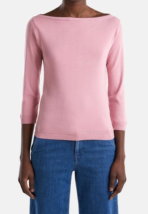 Femme portant un haut rose clair à manches longues et épaules dénudées, associé à un jean bleu taille haute, debout devant un fond uni.