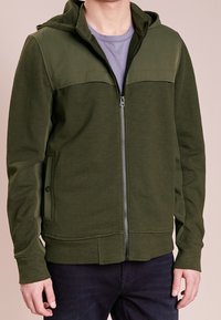 Olivengrøn lynlåshoodie med et dobbeltlagsstofdesign, der har hætte, ribstrikkede manchetter og sidelommer. Glat tekstur.
