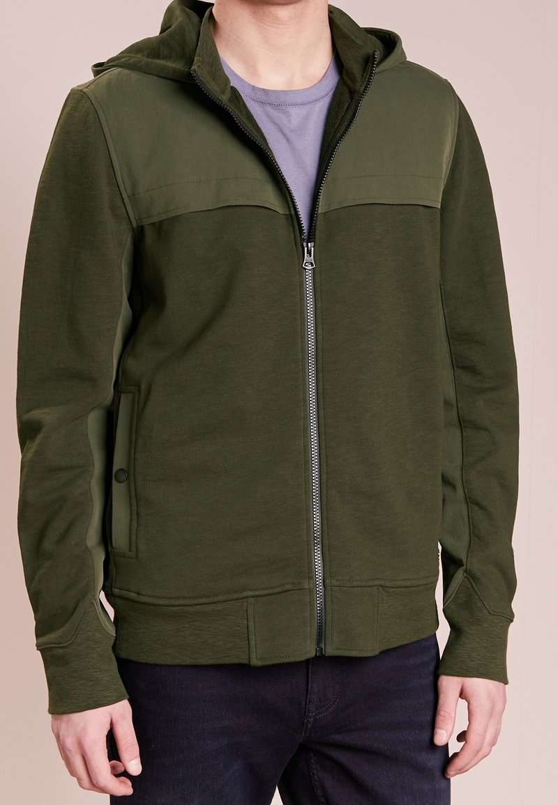 Olivengrøn lynlåshoodie med et dobbeltlagsstofdesign, der har hætte, ribstrikkede manchetter og sidelommer. Glat tekstur.