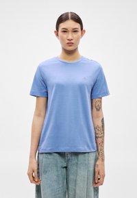 Svetlo modra bombažna majica s prsnim izrezom in kratkimi rokavi. Na prsih je majhen vezen logotip. Tekstura je videti gladka.