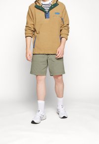 Pullover in pile marrone con accenti verdi, con una chiusura a metà bottone e tasche laterali, abbinato a pantaloni corti verde oliva e scarpe da ginnastica bianche.