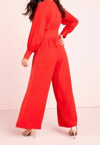 Rode jumpsuit met wijde benen, lange mouwen en een aansluitende tailleband; bevat knopen op de manchetten en een gladde stof; gecombineerd met beige hoge hakschoenen.