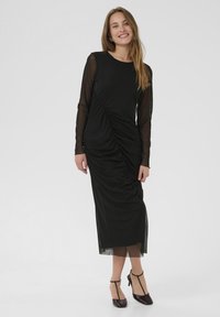 Black dress featuring long sheer sleeves, ruched design down the front, ankle-length with side slits, paired with dark heeled shoes.