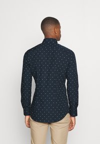 Camicetta blu navy con piccolo motivo geometrico bianco, maniche lunghe e colletto. Vestibilità sartoriale con orlo arrotondato e polsini abbottonati.