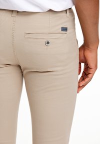 Khaki kalhoty s hladkým povrchem, s kapsou na zadní straně se zapínáním na knoflík a malou značkovou visačkou blízko pasu.