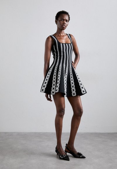 Rochie neagră fără mâneci, cu modele brodate albe, cu o croială ajustată pe corp, fustă evazată și decolteu pătrat. Asortată cu pantofi cu toc ascuțit negri.