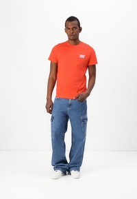 Tricou roșu din bumbac cu logo imprimeriat pe piept, asortat cu pantaloni cargo din denim bleu deschis cu multiple buzunare și adidași albi.