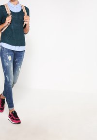 Osoba nosząca niebieską koszulę z kołnierzykiem pod ciemnozielonym swetrem, w zniszczonych ciasnych dżinsach, różowo-czarnych trampkach, niosąca beżowy plecak na ramieniu.