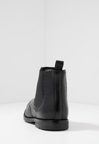 Zwarte leren Chelsea-boot met elastische zijpanelen, brogue-details en een treklus aan de achterkant. Glad oppervlak en platte rubberen zool.