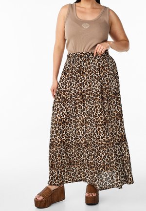 Kobieta w beżowym topie na ramię z motywem muszli, długiej spódnicy w panterkę oraz brązowych plecionych sandałach na platformie, stojąca z ręką na biodrze.