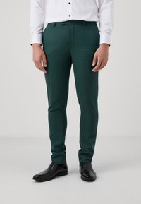 Pantalons vert en tissu lisse, présentés dans une coupe ajustée, avec un devant plat et des poches sur les côtés, associés à des chaussures noires.