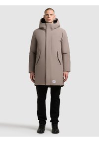 Dlouhá, béžová prošívaná bunda s kapucí, má přední zip, dvě boční kapsy a štítek s logem poblíž lemu. Matný povrch, hladká textura.