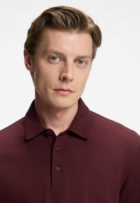 Mörk burgundy polotröja med krage, tre-knappsstängning, textilstruktur och korta ärmar. Modellen har synliga axlar.