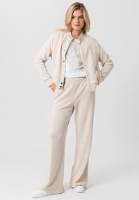 Beige op maat gemaakte jas met drukknoopsluiting, bijpassende hoge broek en een witte t-shirt met grafische opdruk. Gladde textuur, relaxte pasvorm en minimalistisch ontwerp.