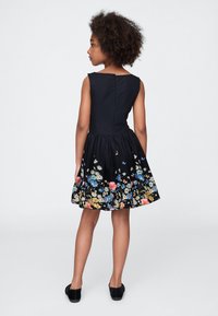 Mergina su garbanotais plaukais, vilkinti juodą rankovių neturinčią suknelę su gėlių raštu apačioje, stovinti ir taikosi nugarą į paprastą foną.