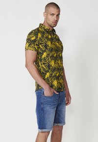 Cămașă cu mânecă scurtă cu un model de frunze tropicale negre și galbene, asociată cu șorturi din denim suflecate. Materialul pare a fi bumbac, textura este netedă.