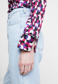 Chemise colorée à motifs géométriques avec des nuances de rose, noir et violet. Elle présente un poignet plié et est associée à un jean en denim bleu clair.