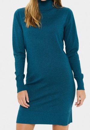 Femme portant une robe-pull à col roulé tricotée en maille teal à manches longues, debout devant un fond uni.
