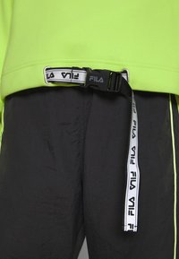 Felpa verde neon brillante con una cintura nera in nylon dotata di una chiusura nera e del logo FILA bianco; pantaloni neri texturizzati con rifiniture gialle.