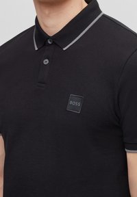 Czarna koszulka polo z teksturowanej tkaniny, z szarym prążkowanym kołnierzykiem, dwoma guzikami oraz kwadratowym, wytłoczonym logo po lewej stronie klatki piersiowej.