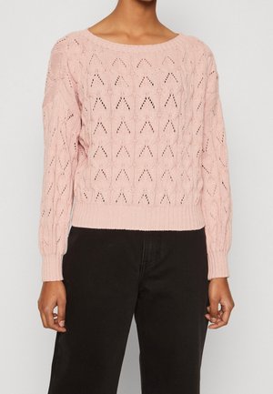 Femme portant un pull en tricot rose clair avec un motif ajouré en forme de cœur et un pantalon noir taille haute.
