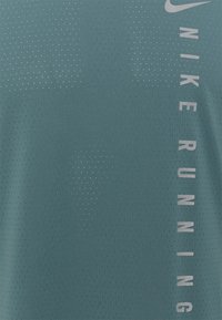 Camiseta de running Nike en verde azulado oscuro, con una tela texturada y transpirable. Presenta un gráfico vertical en gris claro y pequeñas perforaciones en toda la prenda.