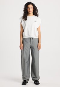 Woman with curly hair wearing a sleeveless white top and loose gray pants, standing against a plain light background.