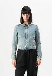 Mlada ženska s pravo črno frizuro, oblečen v svetlo modro zakrpano denim jakno in črne hlače, stoji ob beli ozadju.