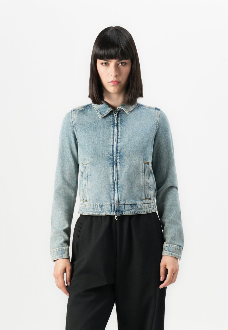 Mlada ženska s pravo črno frizuro, oblečen v svetlo modro zakrpano denim jakno in črne hlače, stoji ob beli ozadju.