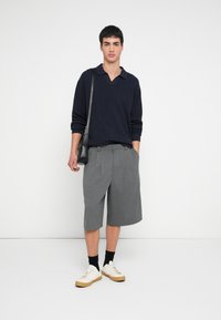 Laivastonsininen neulottu pusero ribbineuloksella, harmaat laskostetut shortsit, musta laukku ja valkoiset lenkkarit vaaleanruskeilla pohjilla yksivärisellä taustalla.