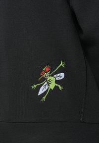 Μαύρο ύφασμα με κεντητό σχέδιο πράσινου εντόμου που διαθέτει κόκκινα μάτια και φτερά, παρουσιάζοντας λεπτομερή ραφή και ζωντανά χρώματα.