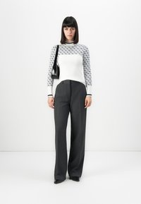 Donna che indossa un maglione bianco a maniche lunghe con motivo, pantaloni larghi grigio scuro, scarpe nere a punta e porta una borsa a tracolla nera.