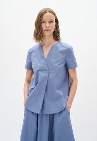 Blusa azzurro chiaro a maniche corte con scollatura a V, caratterizzata da un dettaglio plissettato sul davanti e una vestibilità morbida. Realizzata in un tessuto morbido e testurizzato.
