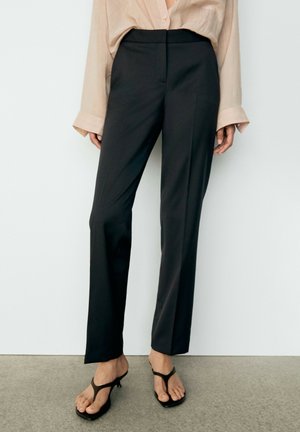 Femme portant un chemisier beige à manches longues rentré dans un pantalon noir taille haute à jambes droites et des sandales à talons noires debout sur un sol gris.