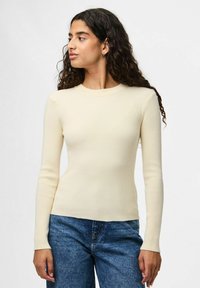 Krämfärgad ribbad långärmad topp med rund halsringning, figurnära design och mjuk textur, i kombination med blå högmidjade jeans.